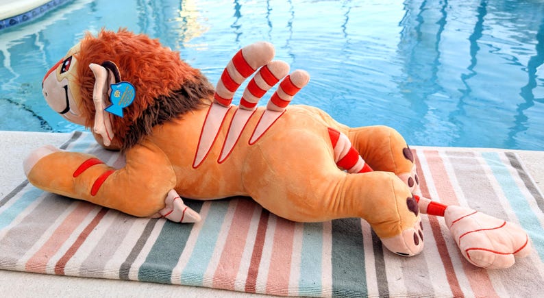 Puede incluir: Un juguete de peluche con forma de le&oacute;n, con pelaje naranja, melena marr&oacute;n y detalles a rayas rojas y blancas. El le&oacute;n tiene una etiqueta azul en la oreja y est&aacute; tumbado en una toalla a rayas cerca de una piscina.