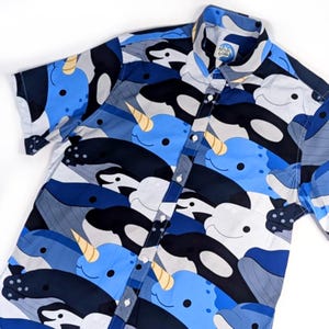 Könnte beinhalten: Ein kurzärmeliges, blau-weiß-schwarzes Hemd mit Knöpfen und einem Narwal- und Wal-Druck. Das Hemd hat eine weiße Knopfleiste und ein kleines Etikett mit dem Text "Whale & Co."