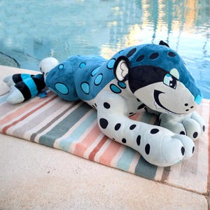 May include: A large plush jaguar toy in shades of blue, black, and white, with black spots and a smiling face. The toy is lying on a striped towel near a pool. The jaguar's body is approximately 48 inches long.