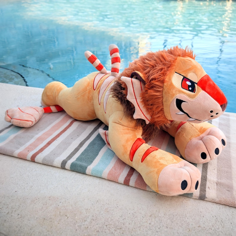 Puede incluir: Un gran peluche de le&oacute;n con un dise&ntilde;o de dibujos animados. El le&oacute;n es principalmente de color beige con detalles en rojo y blanco, incluyendo rayas y detalles en la melena y la cola. Est&aacute; tumbado sobre una toalla a rayas cerca de una piscina.