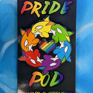 May include: A rainbow pride pin featuring a circle of six orcas surrounding a rainbow heart. The pin says "Pride Pod" in rainbow lettering and "Inner Pin by Artsea Animal" in black lettering.