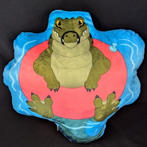 May include: A plush pillow shaped like a crocodile wearing a red inflatable ring, floating on blue water. The crocodile has a toothy grin and is sitting in the ring with its arms crossed.