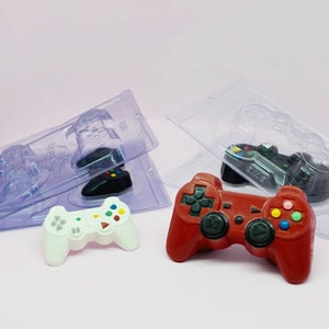 May include: Two plastic molds for making chocolate game controllers. One mold is clear and the other is white. A red and a white chocolate game controller are shown next to the molds.