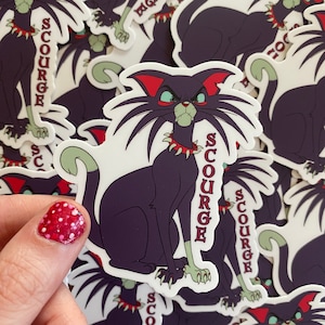 May include: A sticker featuring a cartoon cat with a menacing expression. The cat is dark purple with green accents, a red collar, and the word "SCURGE" printed vertically. The sticker is held by a hand with a red and white polka dot nail design.