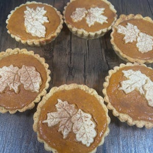 May include: Six mini pumpkin pies with a decorative leaf design. The pies have a golden-brown crust and an orange filling. The leaf design is made of a light-coloured pastry.