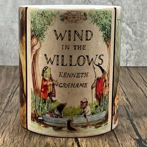 May include: A cylindrical ceramic mug with illustrations from "Wind in the Willows" by Kenneth Grahame. The design features characters in a boat and other scenes from the children's book. The mug has a brown and beige colour scheme.