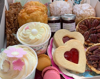 Puede incluir: Una caja blanca llena de una variedad de productos horneados y golosinas. Incluye cupcakes con decoraciones florales, galletas en forma de corazón con mermelada, bollos, tartaletas de nueces y macarons. También contiene bolsitas de té y pequeños frascos de conserva de fresa.