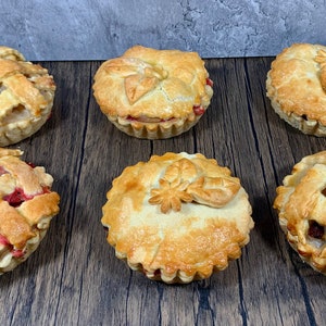 May include: Six mini pies with golden-brown, flaky pastry crusts. Some have lattice tops, others are decorated with leaf-shaped pastry cutouts. The fillings appear to be various fruits, with red and brown hues visible. The pies sit on a dark wood surface.