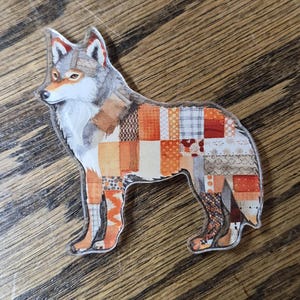 May include: A wolf-shaped acrylic pin featuring a patchwork design across its body. The wolf has a grey and white head with orange accents, while the torso and legs are covered in a grid of various orange, brown, and grey patterned squares.