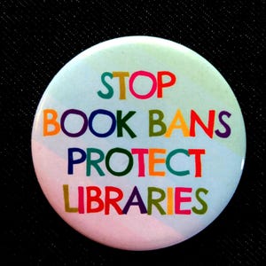 May include: A white button with the text "STOP BOOK BANS PROTECT LIBRARIES" in rainbow colors.