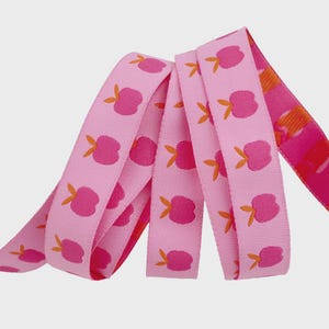 May include: Pink ribbon with a repeating pattern of stylized pink flowers with orange leaves. The ribbon is folded over itself, showing the pattern on both sides. The ribbon is a solid pink color.