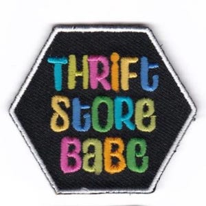 May include: A hexagonal black patch with white stitching around the edges. The text "Thrift Store Babe" is embroidered in colorful letters.