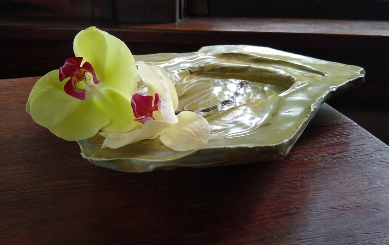 May include: A green ceramic dish with a rough, textured surface. The dish is decorated with a single yellow orchid with a red center.