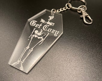 May include: A clear acrylic keychain shaped like a coffin with a skeleton inside and the text "Get Cozy" in a gothic font.