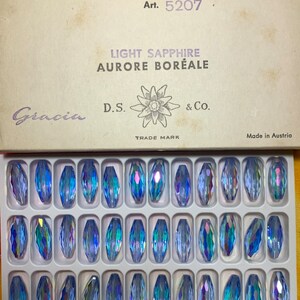 May include: A box of light sapphire aurora borealis crystal beads. The box reads "6/12 gross 17.5 x 7 mm" and "Art. 5207". The beads are a light blue color with iridescent flashes. The box is labeled "Made in Austria".