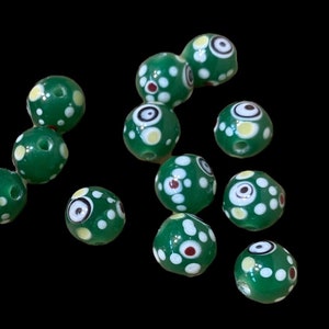 May include: A collection of green glass beads with white, yellow, and red dot patterns. Some beads feature an eye-like design. The beads are round and have a hole for stringing. The background is black.