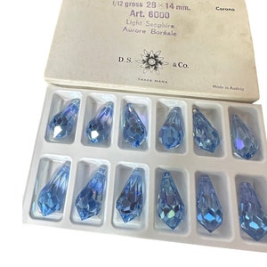 May include: A box of light blue crystal teardrop-shaped pendants. The box is labeled with text including "Light Sapphire" and "Aurore Boréale". Each pendant measures approximately 1.1 x 0.5 inches. The box is marked "Made in Austria".