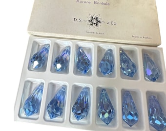 May include: A box of light blue crystal teardrop-shaped pendants. The box is labeled with text including "Light Sapphire" and "Aurore Boréale". Each pendant measures approximately 1.1 x 0.5 inches. The box is marked "Made in Austria".