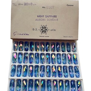 May include: A box of light blue, iridescent crystal rhinestones, each measuring 20 x 8 mm. The box is labeled "Light Sapphire Aurore Boreale" and "Art. 5207". The crystals are arranged in rows within a white tray.