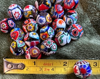May include: A collection of colorful, multi-faceted glass beads with intricate patterns. The beads are approximately 1 inch in diameter and are arranged in a pile on a green fabric surface. A yellow measuring tape is visible in the foreground.
