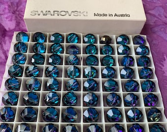 May include: A box of Bermuda Blue Swarovski crystals, 14 mm in size, arranged in a grid. The box is labeled with "Swarovski Made in Austria". The crystals are a deep blue with iridescent highlights, reflecting light.