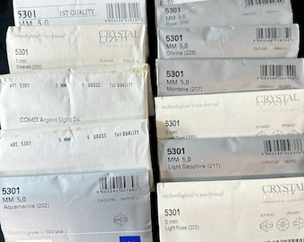 May include: A close-up of a variety of small, clear crystal rhinestones packaged in individual boxes. Each box features a different color and name of the crystal, such as Light Amethyst, Jonquil, Ruby, Rose, Olivine, Montana, Light Sapphire, and Light Rose. The boxes are labeled with the size of the crystals, 5 mm, and the quantity, 5,000 gross = 720 pieces.