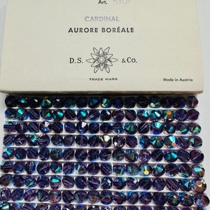 Puede incluir: Primer plano de una caja de cuentas de cristal Cardinal Aurora Boreale de 8 mm. La caja es blanca con texto negro y un logotipo de flor. Las cuentas son de un azul profundo con un brillo iridiscente.
