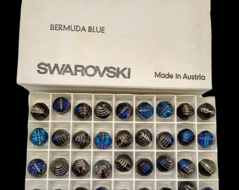 May include: A box of Swarovski Bermuda Blue beads, each 12 mm in diameter, arranged in a grid. The beads have a metallic sheen with blue and iridescent accents. The box is labeled with "SWAROVSKI" and "Made in Austria."
