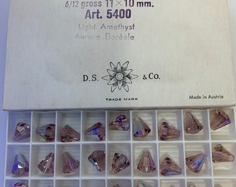 May include: A box of 6/12 gross 11 x 10 mm light amethyst Aurora Borealis crystal beads. The box is labeled "Art. 5400" and "Corona" and is made in Austria. The beads are arranged in a grid in the box.
