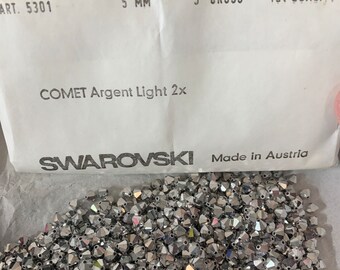 Op de afbeelding: Een stapel zilverkleurige Swarovski-kristallen, met het label "COMET Argent Light 2x", met een afmeting van 5 mm. De verpakking vermeldt "Made in Austria" en "1st QUALITY".