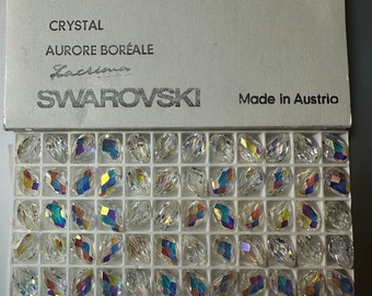 May include: A box of Swarovski crystals, each measuring 12 x 8 mm, with an iridescent finish. The box is labeled "Crystal Aurore Boréale" and "Made in Austria". The crystals are arranged in a grid pattern within the box.