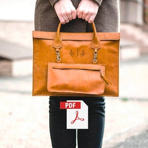 May include: A tan leather tote bag with a zippered pocket and a monogrammed initial "J" on the front. The bag is being held by a person wearing black jeans and white sneakers.