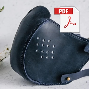 Pode incluir: Uma máscara facial de couro azul escuro com um design costurado e uma fileira de pequenos furos. A máscara tem tiras ajustáveis com fivelas de latão. A imagem inclui o texto "PDF".
