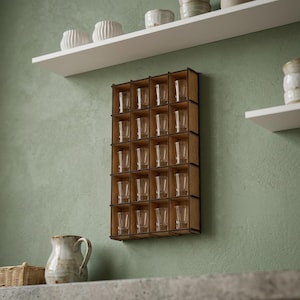 Pode incluir: Um expositor de madeira para copos de shot com 20 compartimentos, cada um com um copo de shot transparente. O expositor está montado numa parede verde. Prateleiras brancas com objetos decorativos estão acima.