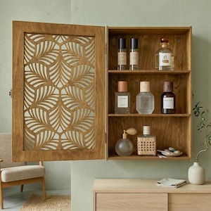 May include: Wooden wall-mounted cabinet with an open door revealing shelves filled with perfume bottles. The cabinet door features a decorative leaf-patterned cutout. The cabinet is made of light brown wood and is mounted on a light green wall.