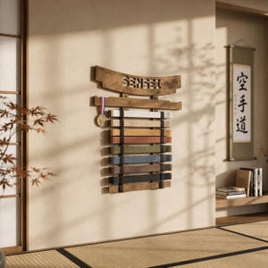 以下が含まれることがあります： 上部に「SENSEI」の文字が切り抜かれた木製の武道帯ディスプレイ。さまざまな色の帯とメダルを保持しています。背景には伝統的な日本の部屋の設定が含まれています。