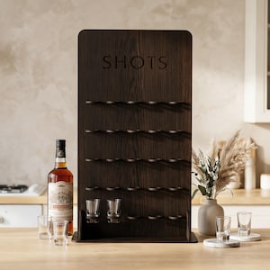 以下が含まれることがあります： 「SHOTS」の文字が刻まれた、濃い茶色の木製ショットグラスディスプレイラック。ラックにはショットグラス用の棚が複数あります。カウンターにはお酒のボトルとショットグラスが置かれています。