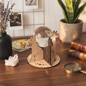 Op de afbeelding: Een heldere kristallen bol staat in een houten katvormige standaard met een cirkelvormige basis. De standaard heeft een lichte houten afwerking met geëtste maanfasen en een oogsymbool. Tarotkaarten en een vergrootglas zijn zichtbaar.