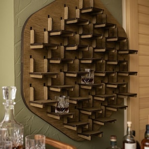Op de afbeelding: Een houten wanddisplay met meerdere kleine planken gerangschikt in een geometrisch patroon. De plank is gemaakt van licht en donkerbruin hout. Twee glazen met een donkere vloeistof staan op de planken. De plank is ontworpen om glazen of kleine items te bevatten.