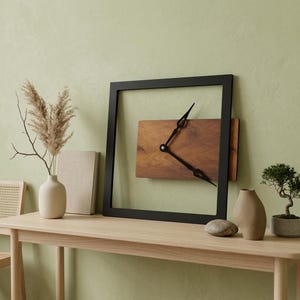 May include: A square wall clock featuring a thin black frame and a rectangular wooden centre panel with visible grain. The clock has two black hands positioned in the middle of the open frame, creating a floating effect against the wall surface.