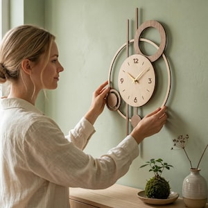 May include: A wall clock featuring a light-colored circular face with gold hands and numerals 3, 6, 9, and 12. The clock is framed by dark wood geometric elements, including two large rings and vertical bars, creating an abstract layered design.