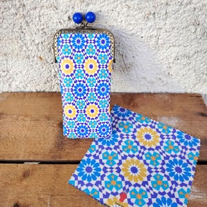 Puede incluir: Un estuche para gafas estampado con un paño de limpieza a juego. El estuche tiene un marco de metal y detalles esféricos azules. El estampado presenta formas geométricas azules, amarillas y blancas. El estuche está sobre una superficie de madera.