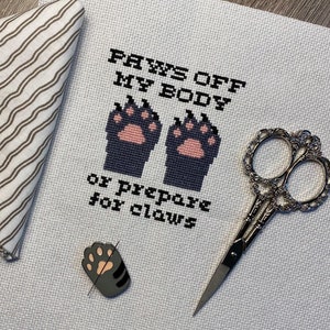 Puede incluir: Bordado de punto de cruz en tela Aida blanca con hilo negro. El dise&ntilde;o presenta dos huellas de patas negras con almohadillas rosadas y el texto "PAWS OFF MY BODY or PREPARE for CLAWS".