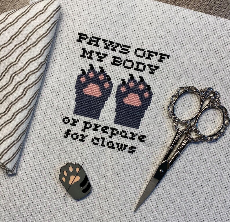 Puede incluir: Bordado de punto de cruz en tela Aida blanca con hilo negro. El dise&ntilde;o presenta dos huellas de patas negras con almohadillas rosadas y el texto "PAWS OFF MY BODY or PREPARE for CLAWS".