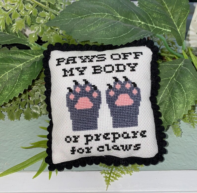 Puede incluir: Una almohada blanca con ribete negro y texto bordado en negro que dice "PAWS OFF MY BODY or prepare for claws". La almohada presenta dos huellas de patas negras con almohadillas rosas.