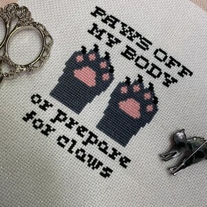 Puede incluir: Bordado blanco de punto de cruz con huellas de patas negras y rosas y el texto "PAWS OFF MY BODY or prepare for claws".