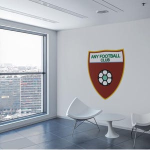 Op de afbeelding: Een witte muur met een rood en groen voetbalclublogo met de tekst "ANY FOOTBALL CLUB". Er staan twee witte stoelen en een witte tafel voor de muur.