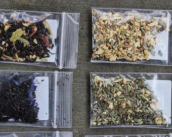 May include: An overhead shot shows several small, clear plastic bags filled with various loose-leaf teas. The teas display a range of colors, including dark brown, green, and hints of blue. Each bag is sealed and contains a different blend of tea.