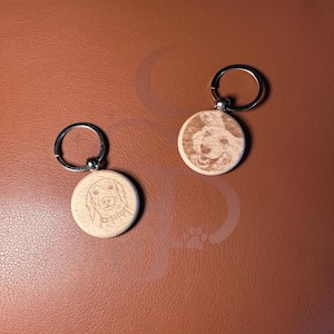 May include: Two circular light-coloured wooden keychains featuring laser-engraved portraits of dogs. Each keychain is attached to a silver-toned metal split ring. The engravings show detailed facial features of the dogs, including eyes, noses, and ears.