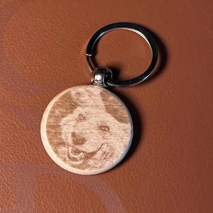 May include: A round wooden keyring featuring a laser-engraved portrait of a dog. The light-coloured wood disc is attached to a circular silver-toned metal split ring. The engraving shows the detailed facial features of a canine with a smiling expression.
